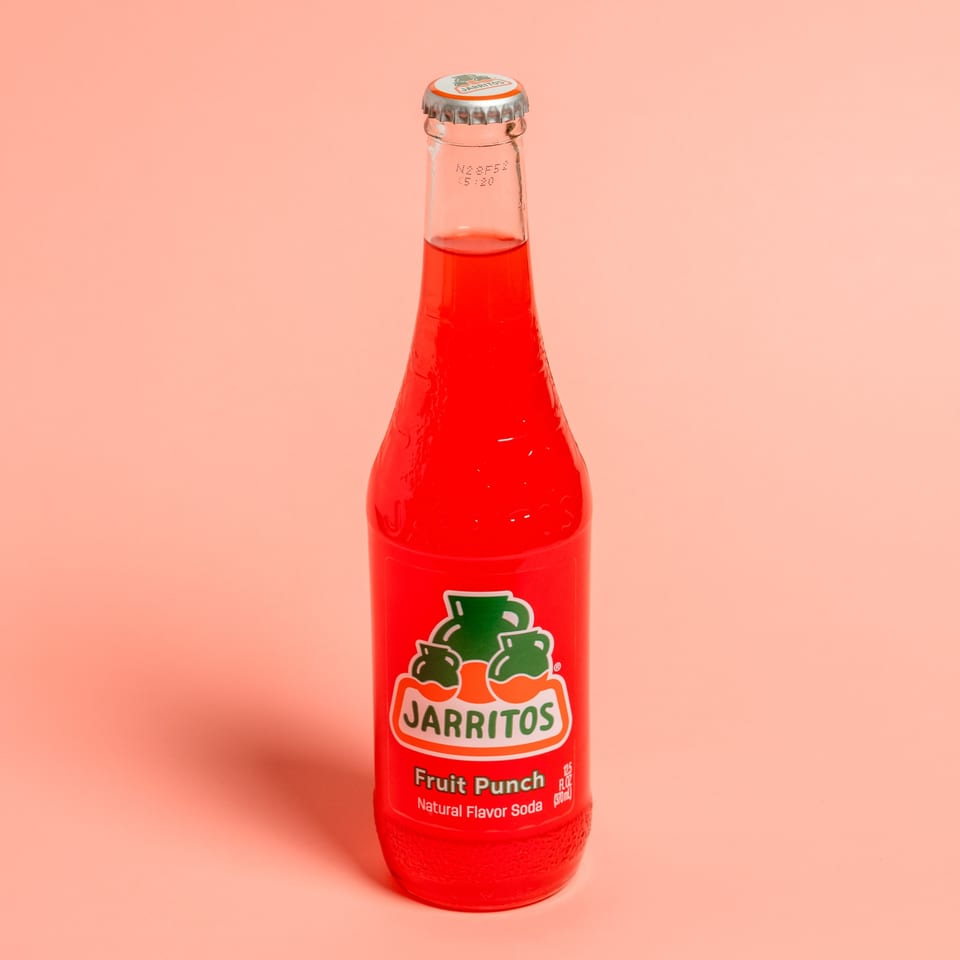 A bottle of red Jarritos Fruit Punch soda stands against a peach background.